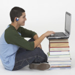 Teen boy with headphones listens to accessible books read aloud on his computer while following along with the text.  His computer is piled high on top of printed books.