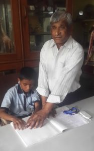 Suraj, a student in Pune, India, reads a braille book while listening to the narration on a Plextalk player with help from Dr. Homiyar.