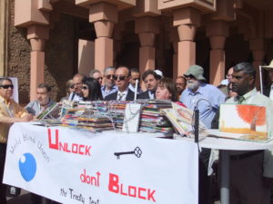 Group of people stand behind a table holding books covered in chains and banner saying "Unlock, don't block"