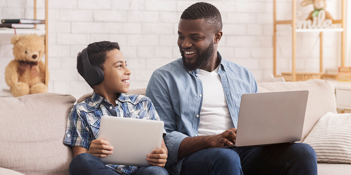 A boy with headphones and a tablet is sitting on a couch next to his father who is using a laptop