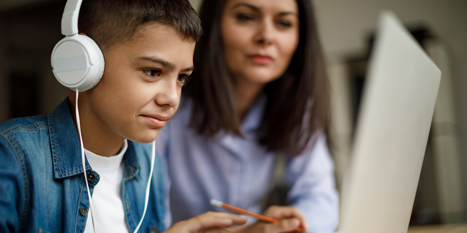A young boy wears headphones and uses a computer with his mother sitting next to him
